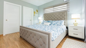 Une chambre à coucher avec une tête de lit capitonnée, un lit recouvert d’une couvre-lit à motifs floraux, une table de chevet avec une lampe et une fenêtre avec des stores.