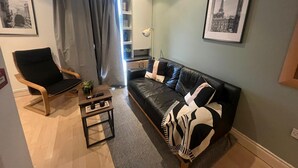 A living room with a black leather sofa, a wooden chair, a small table with a plant, and a grey curtain. - Cozy flat in Belfast (Belfast)
