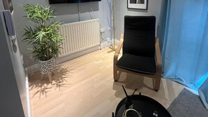 A modern living room with a flat-screen TV mounted on the wall, a black chair, a small table with a remote, and a potted plant. - Cozy flat in Belfast (Belfast)