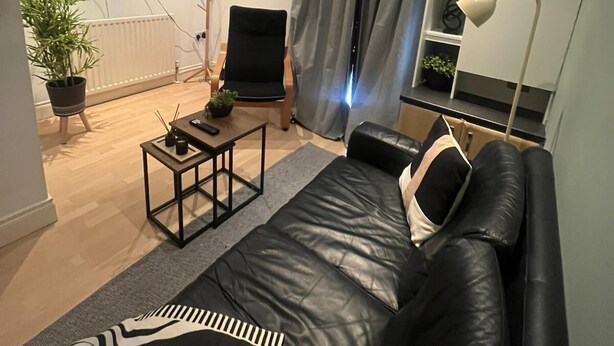 A modern living room with a black leather sofa, a small coffee table, a black chair, a TV mounted on the wall, a potted plant, and a grey curtain. - Cozy flat in Belfast (Belfast)