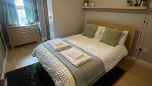 A bedroom with a bed, a bedside table, a nightstand, a window with curtains, a ceiling light, and a shelf with decorative items. - Cozy flat in Belfast (Belfast)