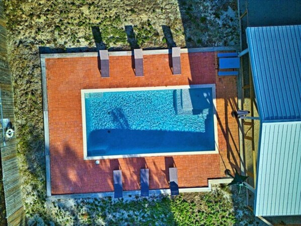 Vue aérienne d’une piscine rectangulaire bordée d’une terrasse en briques, entourée d’une clôture en bois et surmontée d’une structure de toit en métal.