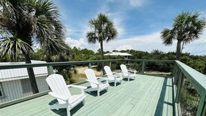 A wooden deck with white Adirondack chairs and a view of palm trees and a building in the background. - Quads-For-Fun, Unit 4, Saint George Island, FL (Eastpoint)