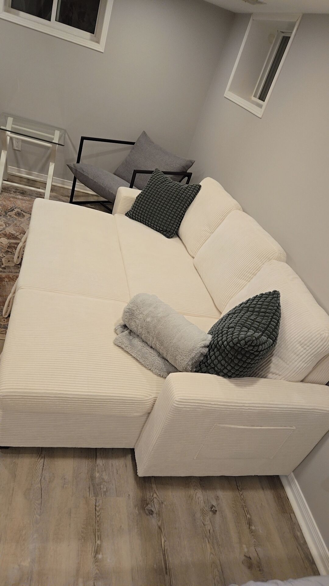 A beige sectional sofa with a grey throw blanket and two grey pillows in a room with wooden flooring and a white wall.