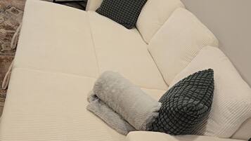 A beige sectional sofa with a grey throw blanket and two grey pillows in a room with wooden flooring and a white wall.