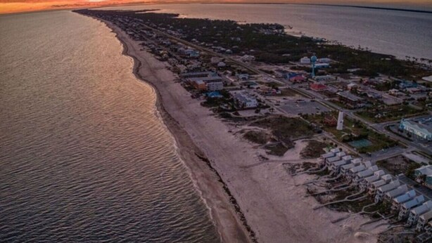 Vista aérea de un pueblo costero con una extensa playa, edificios y una carretera que bordea la orilla.