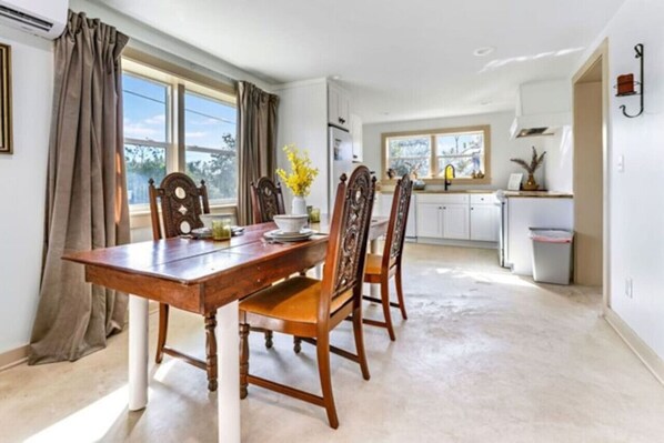 A dining area with a wooden table and chairs, a window with curtains, and a kitchen in the background. - Quads-For-Fun, Unit-1 Saint George Island, FL (Eastpoint)