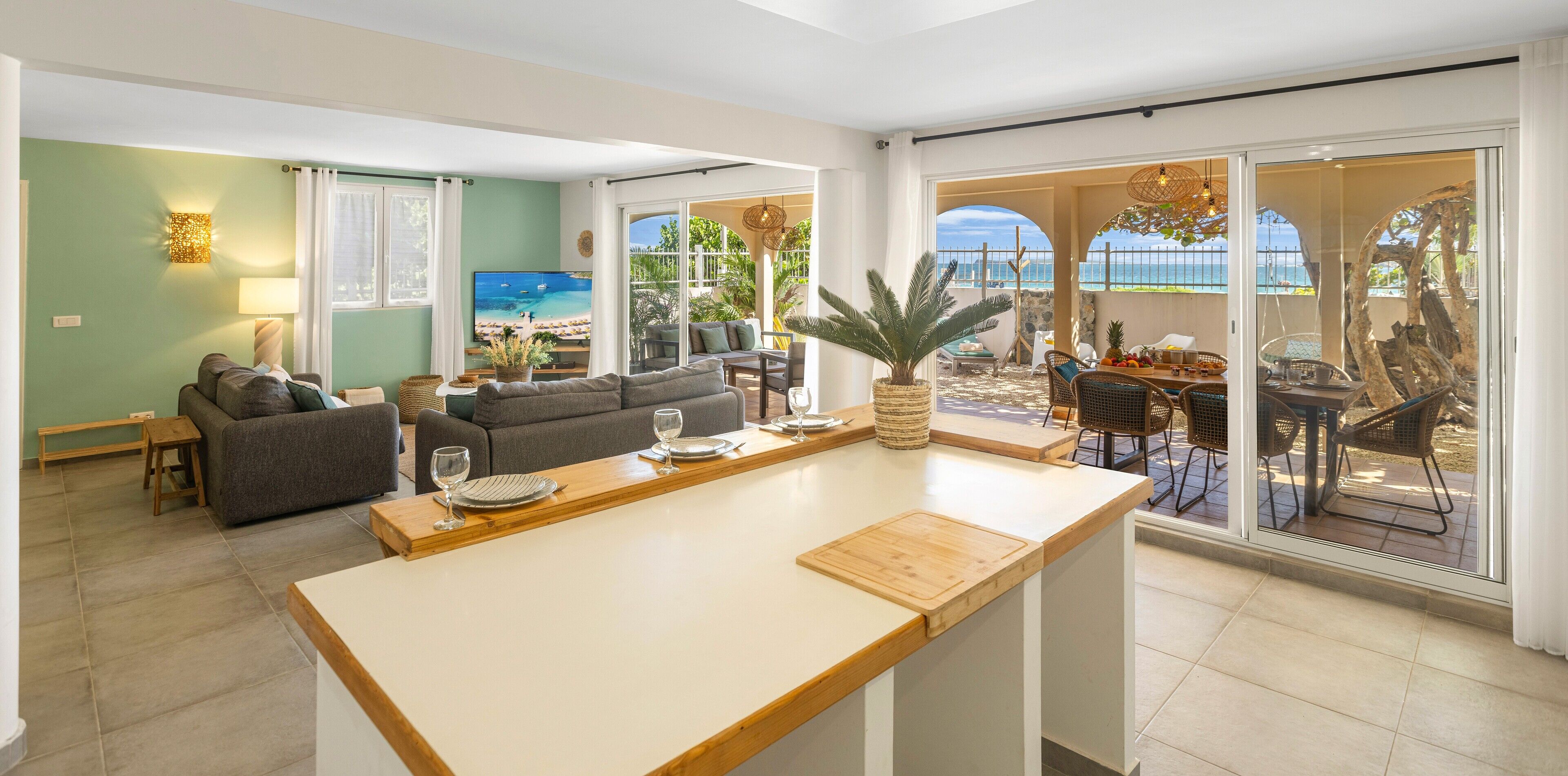 Ein modernes Wohnzimmer mit einem großen Fernseher, einem Essbereich mit einem Holztisch und Blick auf den Strand durch große Schiebetüren.
