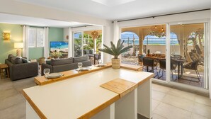 A modern living room with a large TV, a dining area with a wooden table, and a view of the beach through large sliding doors.