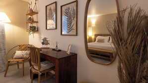 A cozy bedroom with a wooden desk, a chair, a bed, a large mirror, and wall-mounted shelves with framed botanical prints.