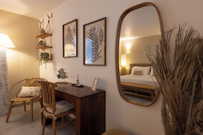 A cozy bedroom with a wooden desk, a chair, a bed, a large mirror, and wall-mounted shelves with framed botanical prints.