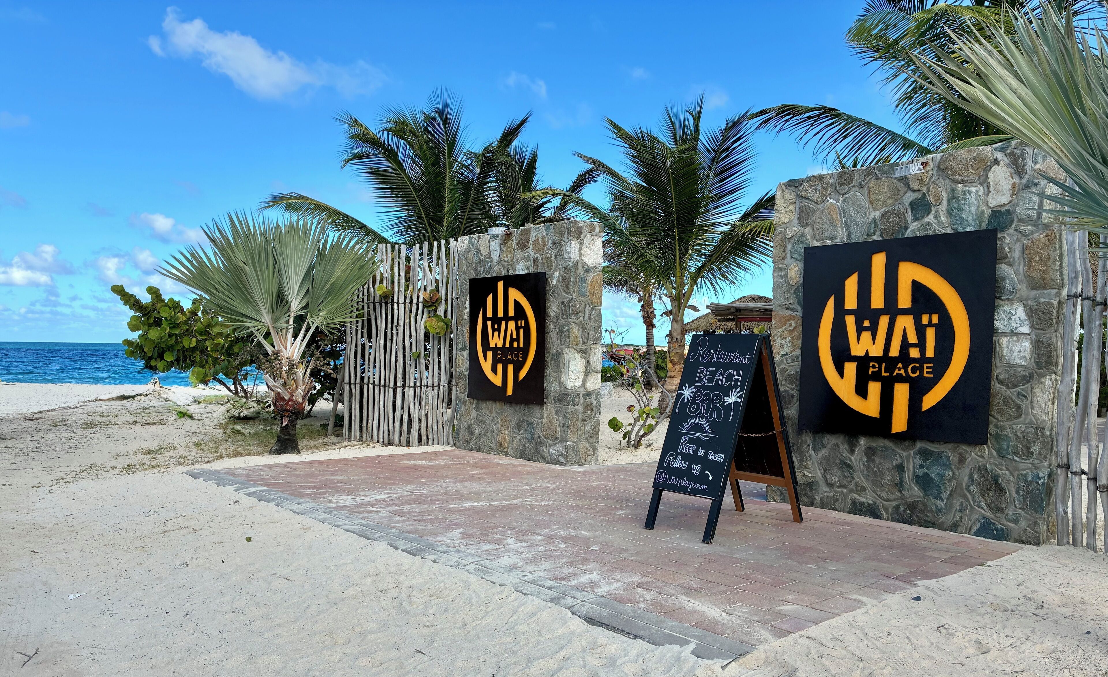 A beachside restaurant entrance with a stone wall, palm trees, and a clear view of the ocean.