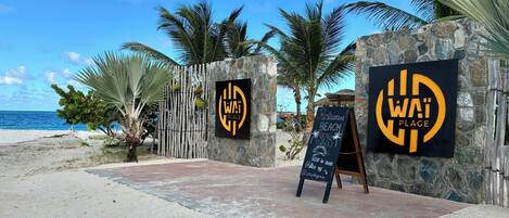 A beachside restaurant entrance with a stone wall, palm trees, and a clear view of the ocean.