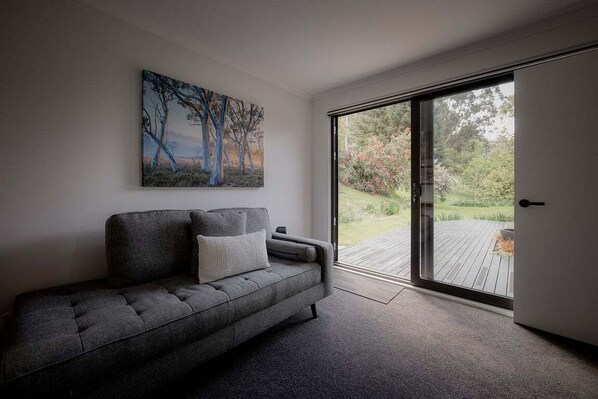 A modern living room with a grey sofa, a painting on the wall, and a sliding glass door leading to a wooden deck. - Wilder Stay with sauna, outdoor bath and plunge (Hobart City)