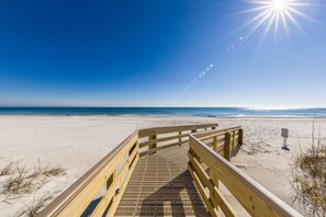 Ein hölzerner Steg führt zu einem Strand mit blauem Himmel und Sonnenschein.