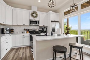 Una cocina moderna con armarios blancos, una isla central, microondas, cafetera y vistas al exterior a través de grandes ventanales.