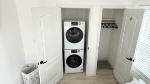 A laundry room with two stacked washing machines, a trash can, and a door leading to a closet with a hanging rack. - Private and charming Bay Area home. Centrally located to all Bay Area cities! (South San Francisco)