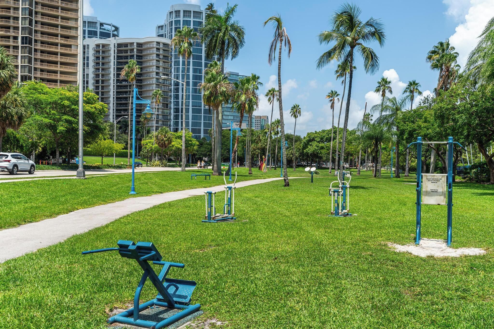 A park with exercise equipment, a paved path, and palm trees.