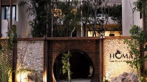 Uma entrada em arco de pedra com a placa "HOMA" iluminada, rodeada por vegetação e um caminho de areia.