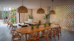 Um espaço amplo e interno com uma mesa de jantar de madeira cercada por cadeiras, um sofá e duas luminárias de vime suspensas.