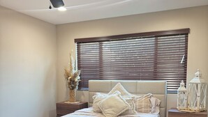 Un dormitorio con cama, ventilador de techo, ventana con persianas, mesita de noche y un farol decorativo.