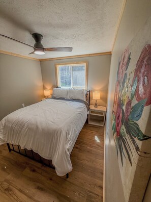 A bedroom with a bed, a ceiling fan, a window, a nightstand, a lamp, and a floral painting on the wall. - Family Retreat | Waterfront | Dock | Sleeps 12 (Hammond)