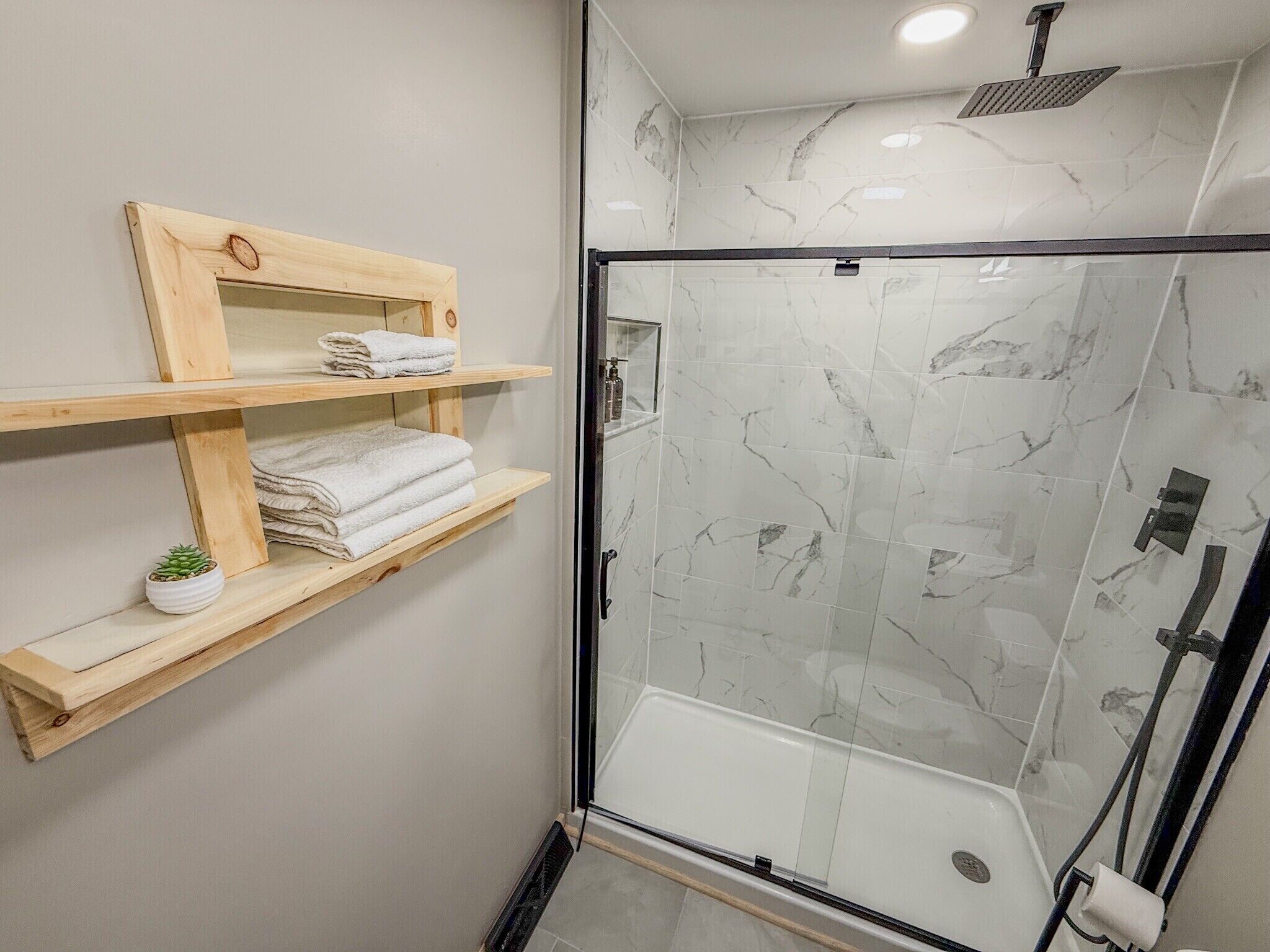 A glass-enclosed shower with marble tiles, wooden shelves with towels, and a small potted plant.