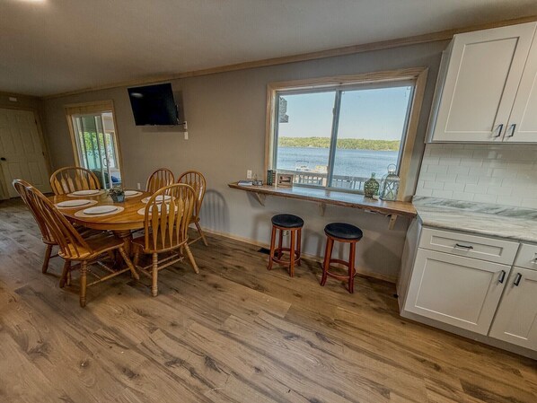 A dining area with a wooden table and chairs, a window with a view of water, and a kitchen with white cabinets. - Family Retreat - Waterfront cabin (Hammond)