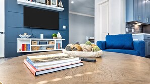 A modern living room with a wooden coffee table, books, a bowl of decorative items, a blue sofa, and a flat-screen TV mounted on a blue wall. - Walk to Palm Beach | Hot Tub + Game Room (Galveston)
