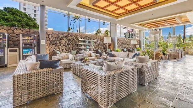 A spacious outdoor lounge area with wicker furniture, a stone wall, and a large window offering a view of palm trees and buildings. - Great View &Waikiki Famliy Retreat (Honolulu)