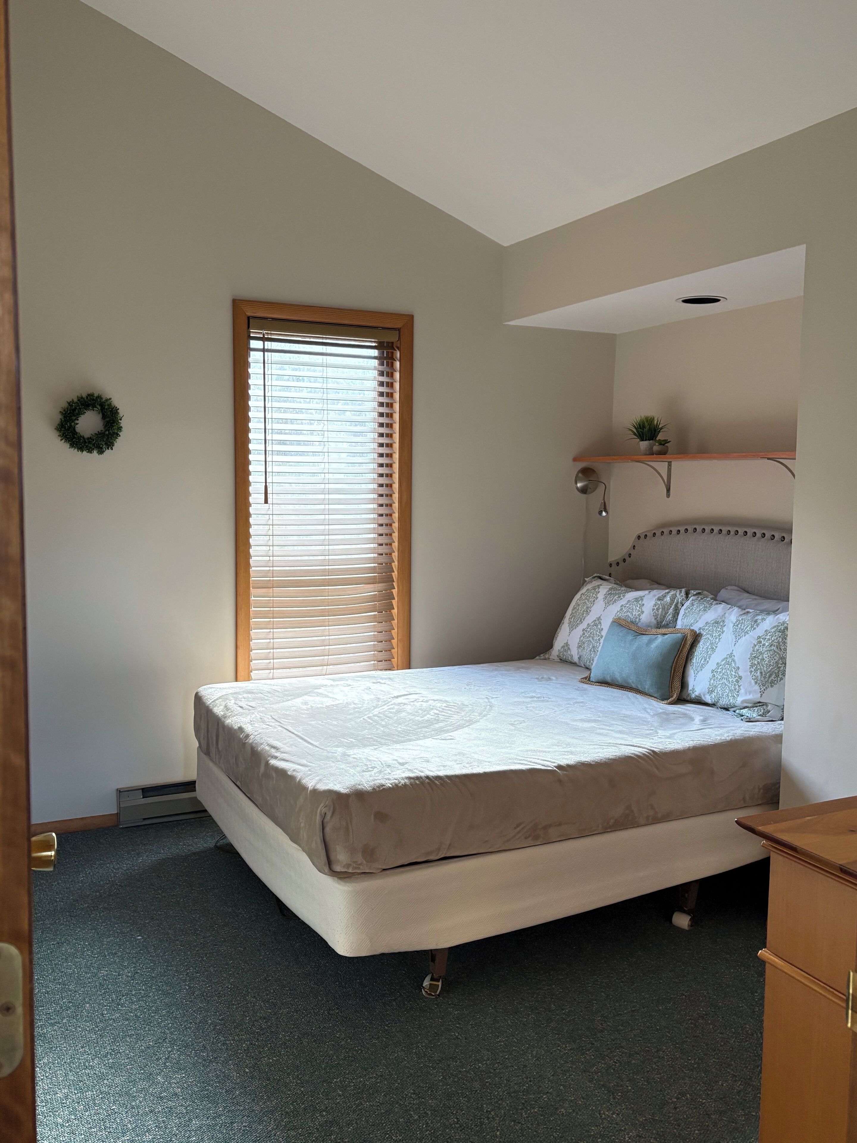 Une chambre à coucher avec un lit, une fenêtre avec des stores, une petite étagère avec une plante et une lampe fixée au mur.
