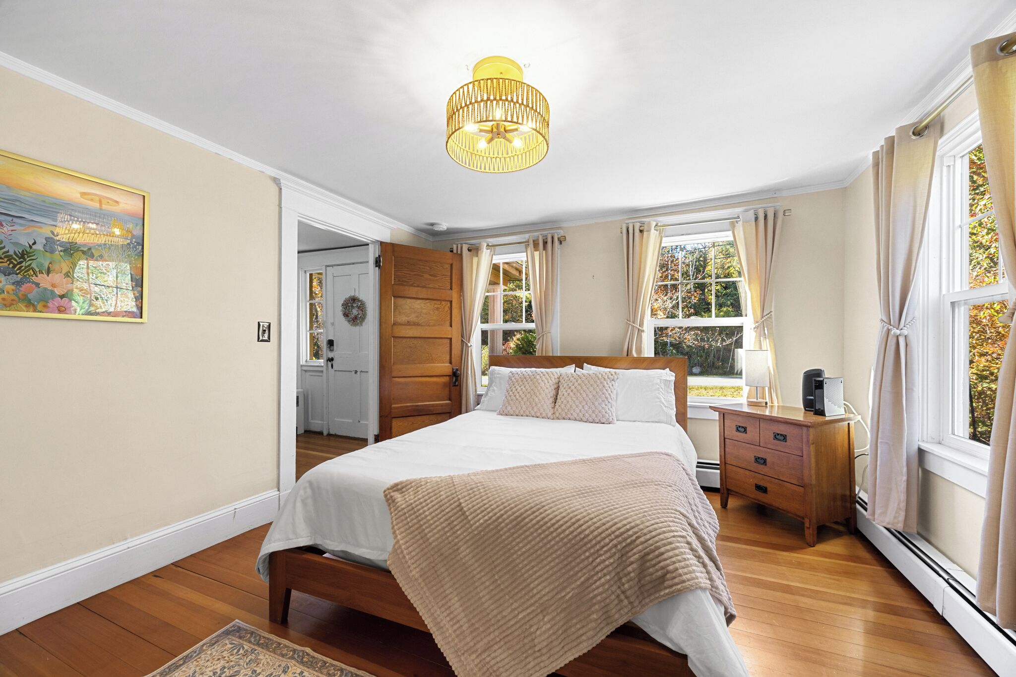 Une chambre avec un grand lit, un plancher en bois, un lustre, une table de chevet et une fenêtre avec des rideaux.