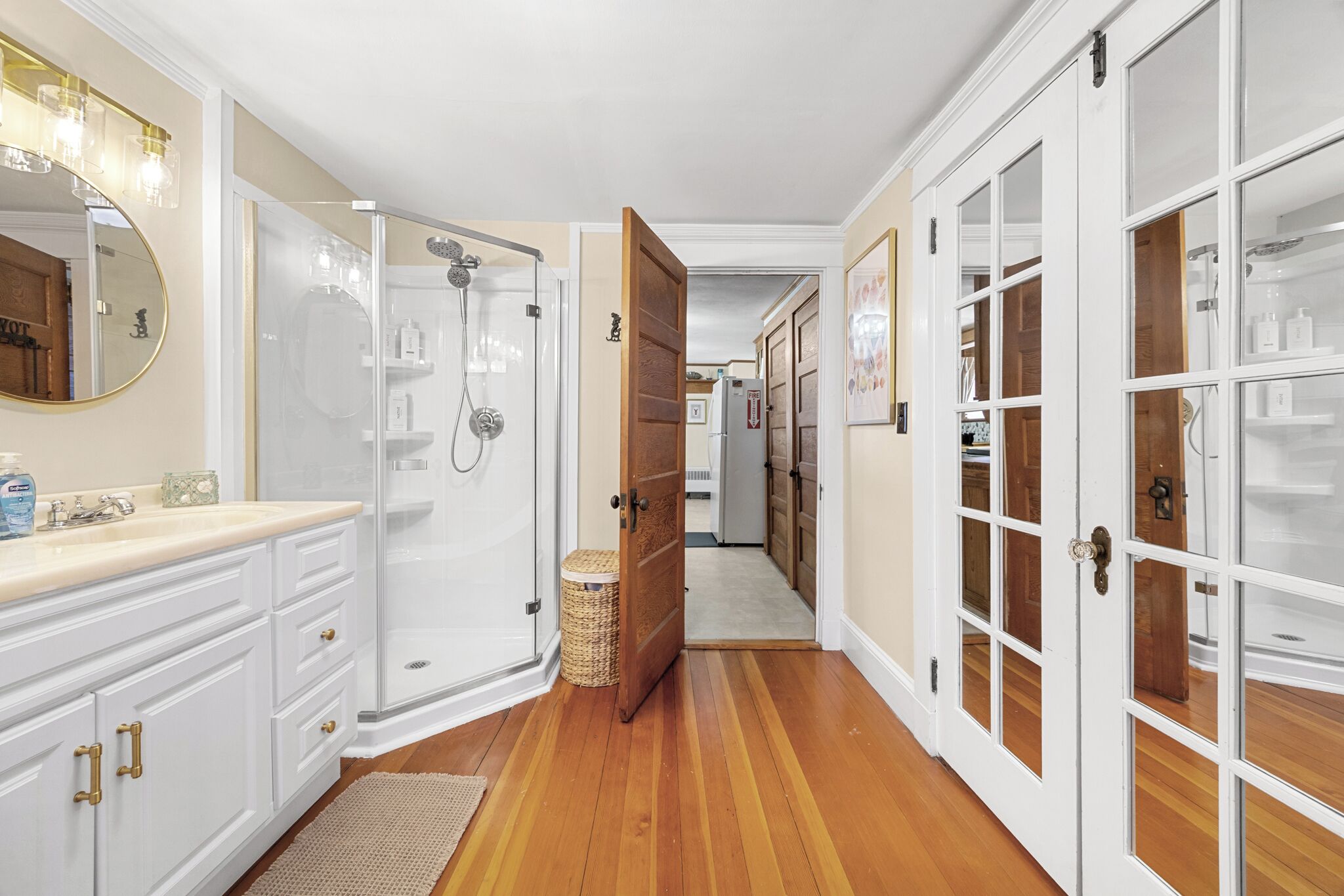 Un couloir avec une salle de bain sur la gauche, comprenant une douche, un meuble-lavabo et un miroir. Des portes en bois se trouvent sur la droite et un panier est posé sur le sol.