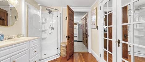 Un couloir avec une salle de bain sur la gauche, comprenant une douche, un meuble-lavabo et un miroir. Des portes en bois se trouvent sur la droite et un panier est posé sur le sol.