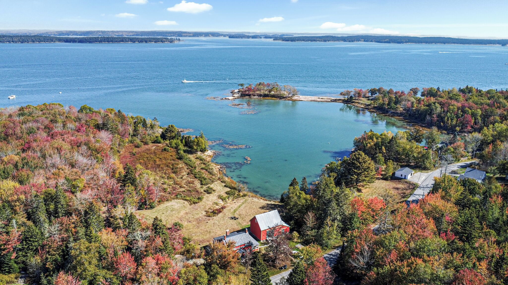 A coastal landscape with a small red building, likely a cabin or lodge, surrounded by dense forest with autumn foliage. A large body of water is visible in the background, with a small island and a boat.