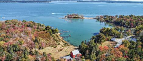 A coastal landscape with a small red building, likely a cabin or lodge, surrounded by dense forest with autumn foliage. A large body of water is visible in the background, with a small island and a boat.