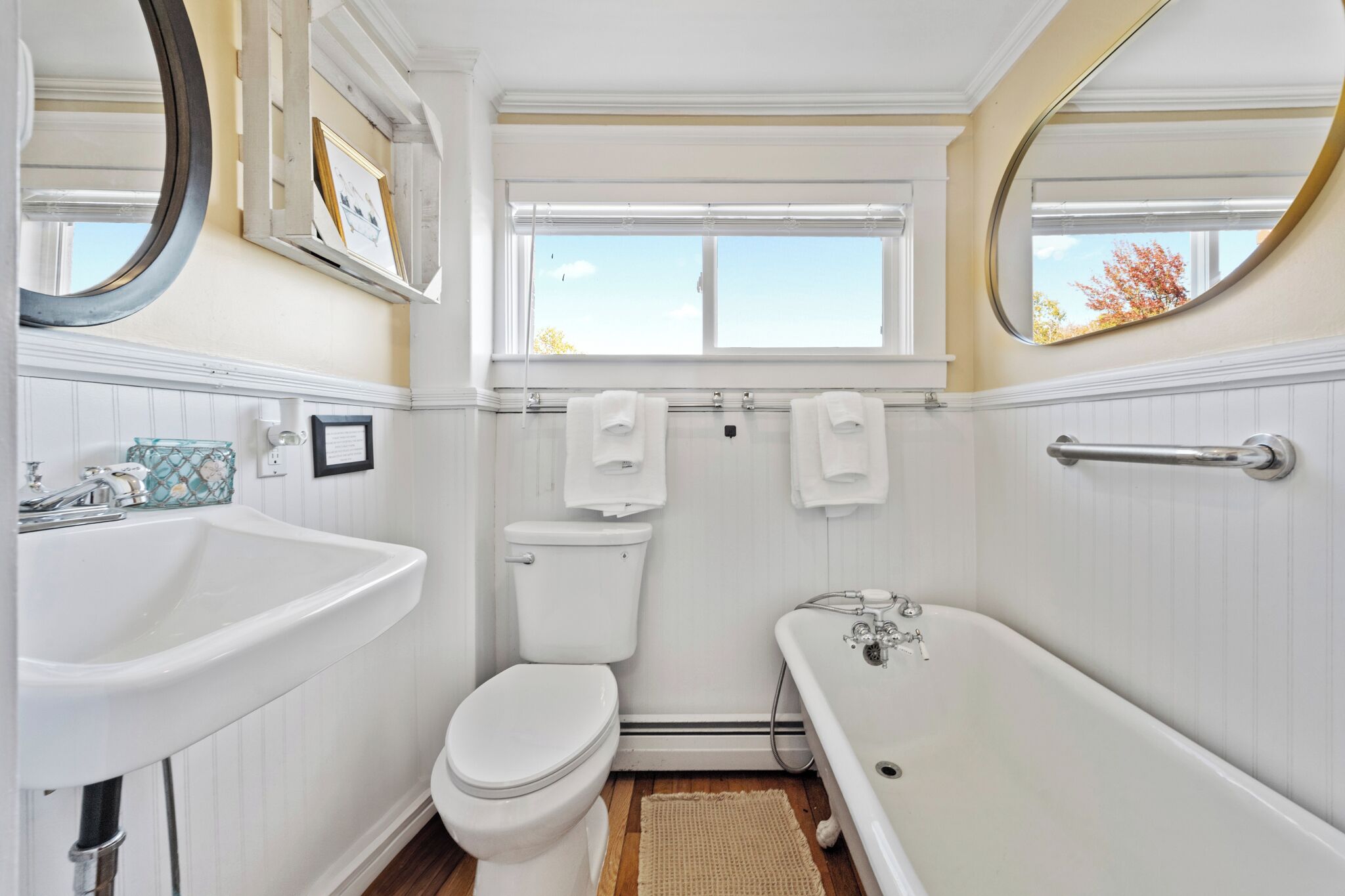 A bathroom with a white sink, toilet, and bathtub, a window with a view of trees, and a mirror above the sink.