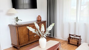 A living room with a wooden coffee table, a white sofa, a TV mounted on the wall, a wooden cabinet, and a window with curtains.