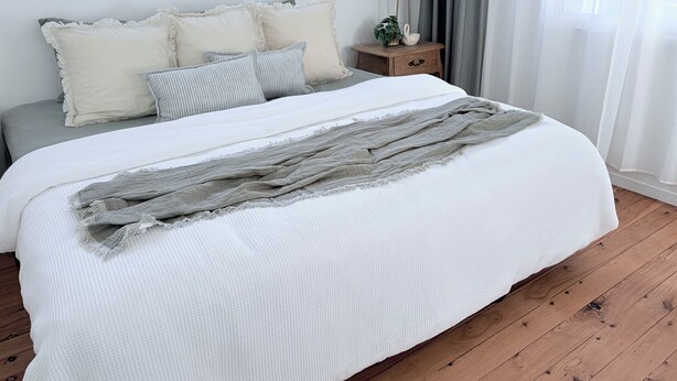 A neatly made bed with a white comforter and a gray throw blanket, a bedside table with a lamp, and a small plant.