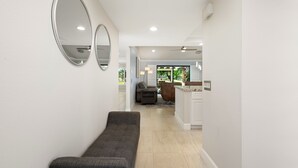 Un interior moderno con un banco, dos espejos en la pared, un ventilador de techo y una cocina visible al fondo.
