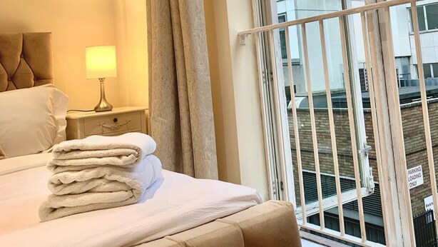 Een hotelkamer met een bed, een nachtkastje met een lamp, een raam met uitzicht op een gebouw en een balkonhek.