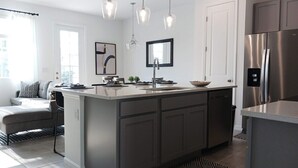 Een moderne keuken met een centraal eiland, roestvrijstalen apparatuur, hanglampen en uitzicht op de woonkamer.