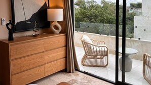 Um quarto moderno com uma cama grande, cômoda de madeira, televisão de tela plana e varanda com móveis de vime e vista para o verde.
