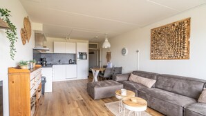 A modern living room with a grey sofa, wooden coffee table, and a kitchen in the background. - 4-person Apartment With Island-sea View (De Koog)