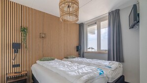 A modern bedroom with a wooden slat wall, a large bed with white bedding, a bedside table with a remote control, and a window with curtains. - 4-person Apartment With Island-sea View (De Koog)