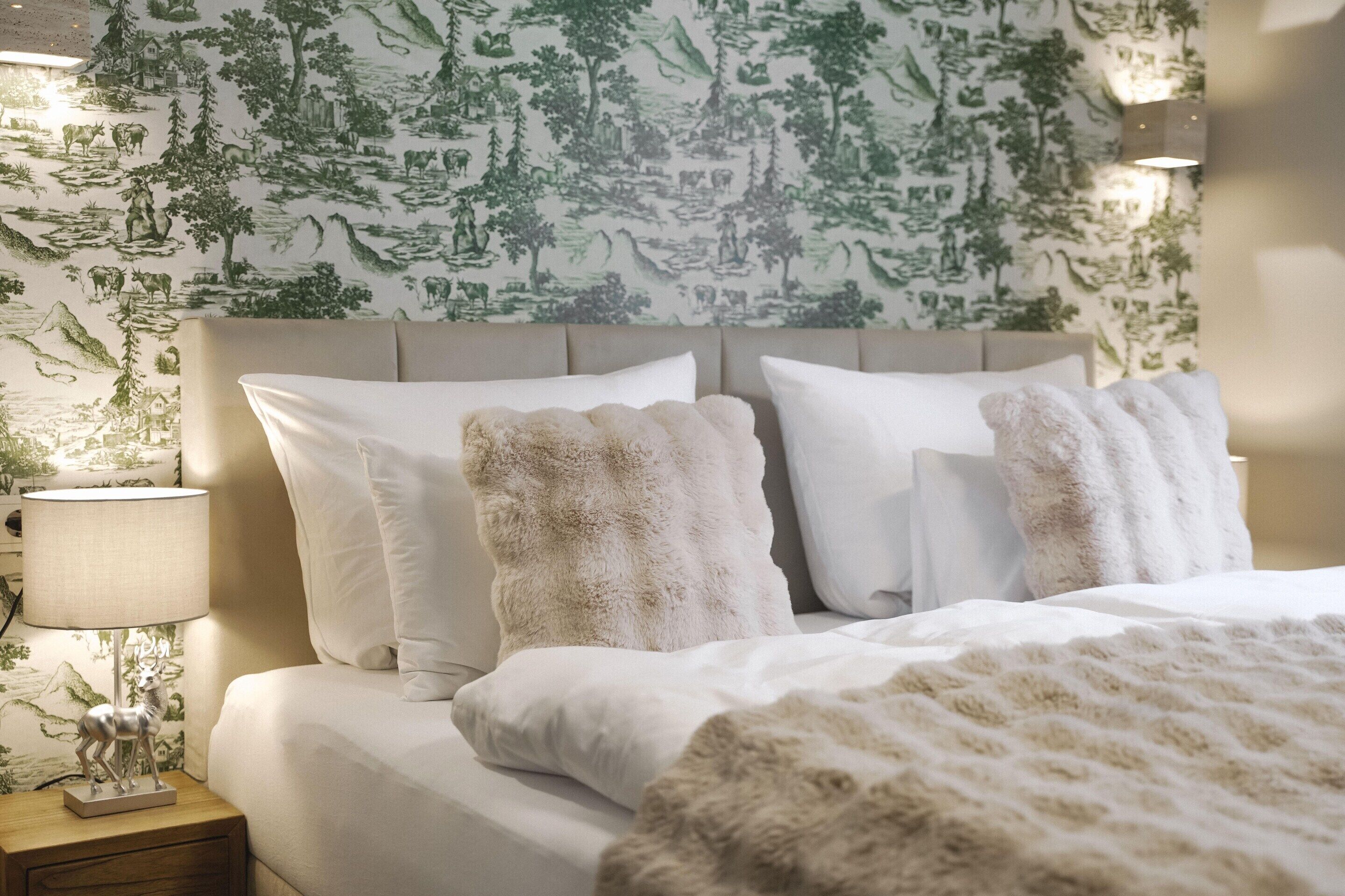 Un lit bien fait, avec du linge de lit blanc et des oreillers moelleux, une lampe de chevet et un mur recouvert d’un papier peint à motifs naturels.