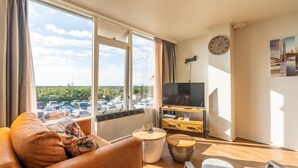 Ein modernes Wohnzimmer mit Sofa, Couchtisch und Fernseher. Ein großes Fenster bietet Blick auf einen Parkplatz und Bäume. An der Wand befinden sich eine Uhr und ein Gemälde.