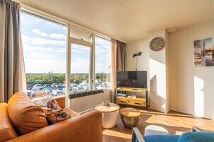 Ein modernes Wohnzimmer mit Sofa, Couchtisch und Fernseher. Ein großes Fenster bietet Blick auf einen Parkplatz und Bäume. An der Wand befinden sich eine Uhr und ein Gemälde.