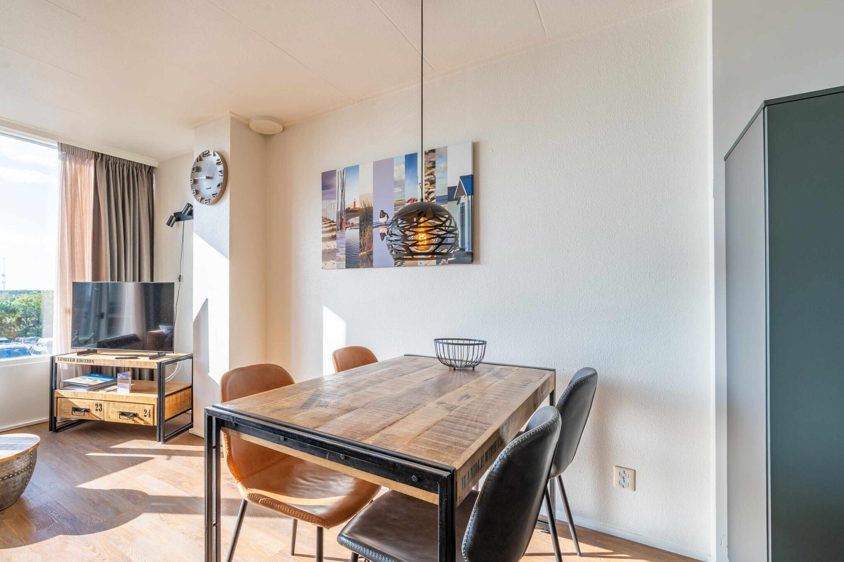 Un salon moderne avec une table à manger en bois, des fauteuils en cuir, un téléviseur à écran plat et une grande fenêtre agrémentée de rideaux.
