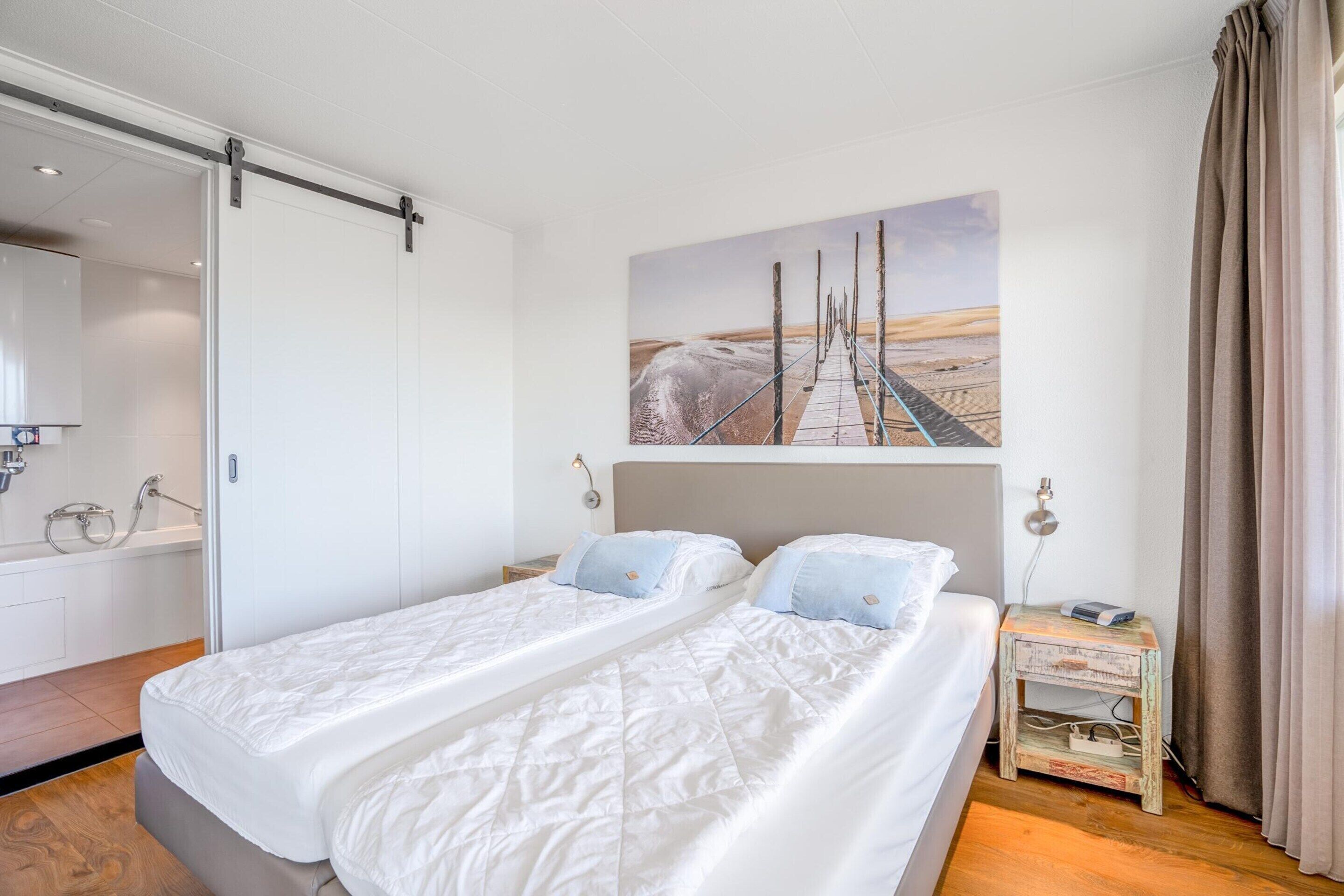A modern hotel room with a large bed, a painting of a beach scene, a sliding door, a small bedside table, and a bathroom visible through the door.